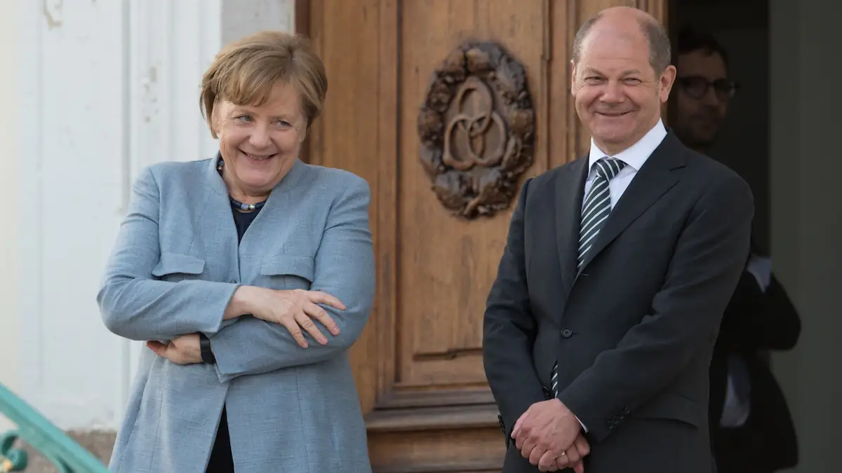 Angela Merkel steht neben Olaf Scholz auf einer Treppe, beide lächeln