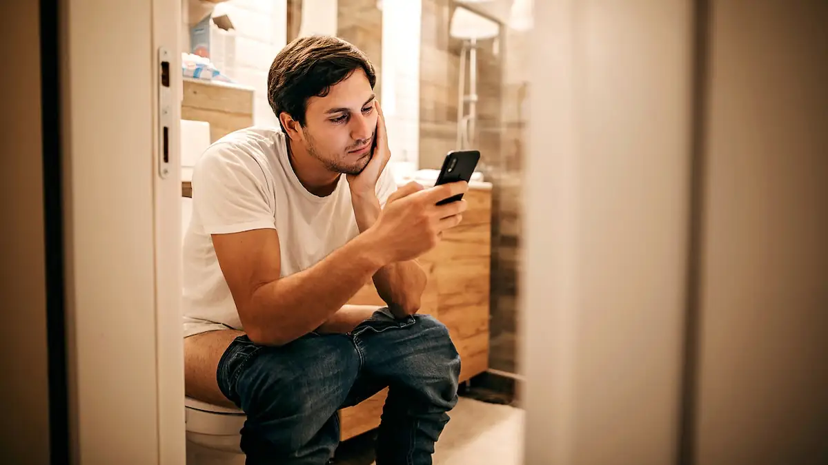 Mann sitzt mit Smartphone auf einer Toilette