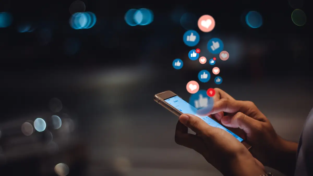Facebook auf dem Smartphone: Jemand hält ein Mobiltelefon in der Hand, über dem diverse Symbole der Interaktion wie Like-Daumen oder Herzen schweben