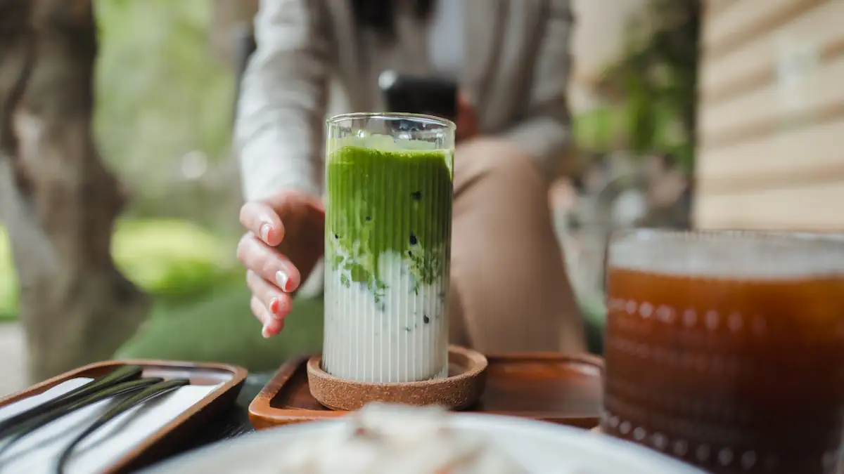 Junge Frau greift nach einem Matcha-Latte-Getränk in einem Glas, während sie es fotografiert