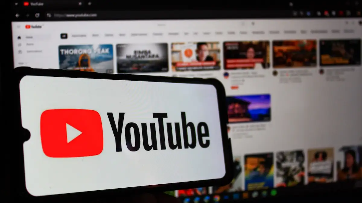 YouTube-Logo auf einem Smartphone, das vor einen Bildschirm gehalten wird, der eine Übersicht der Videoplattform zeigt