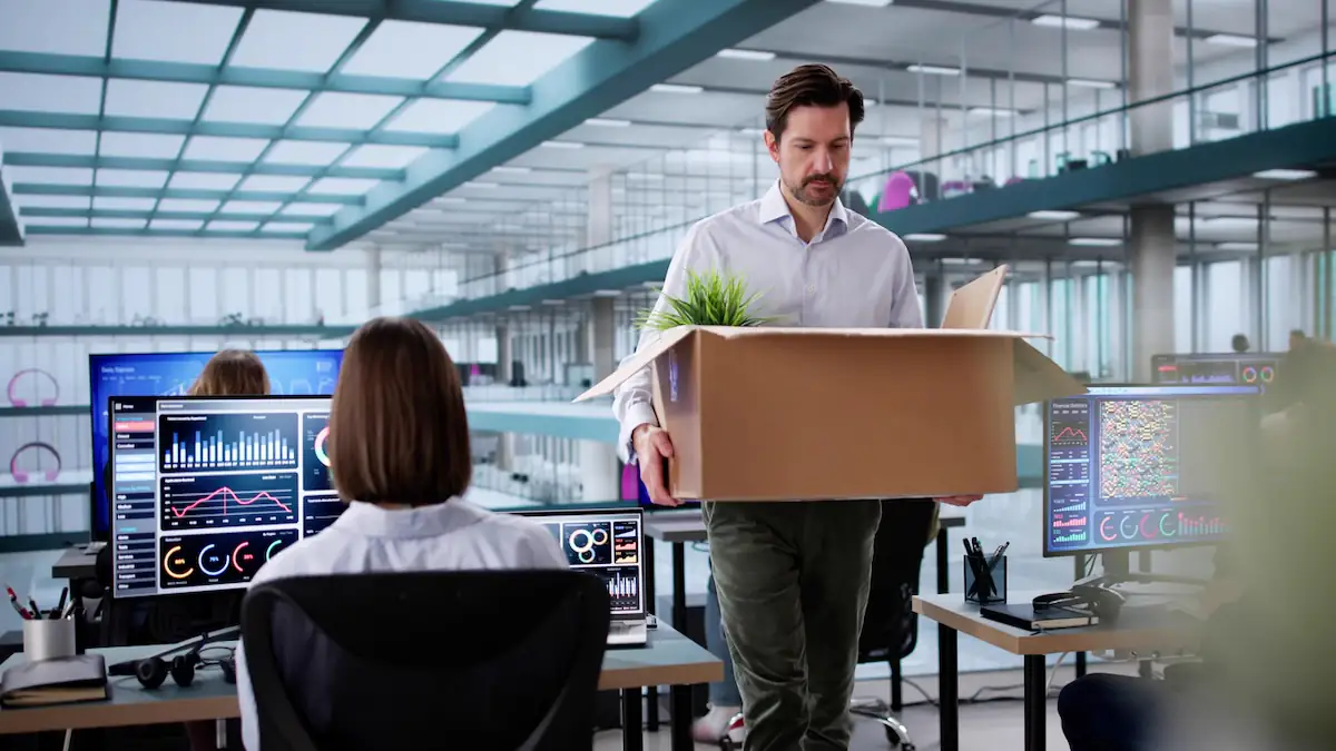 Symbolbild für KI-bedingte Entlassungen: Mann verlässt mit vollem Karton ein Büro, in dem viele Menschen an Computern mit Diagrammen arbeiten