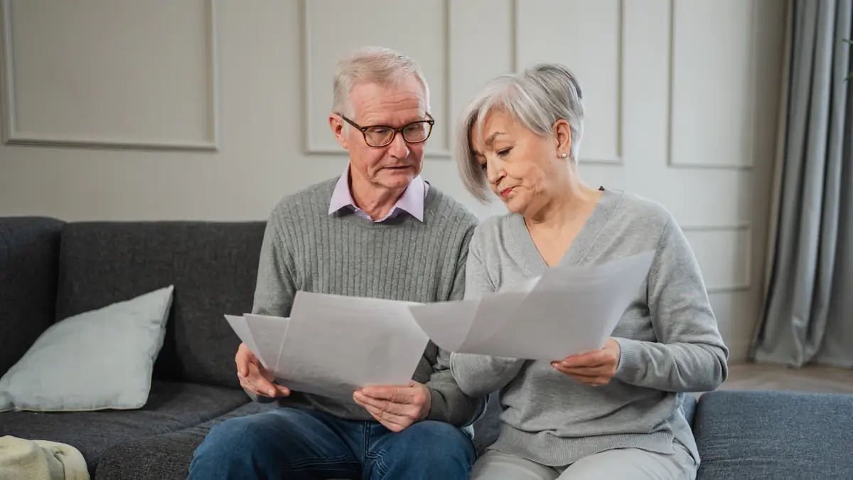 Symbolbild Rente: Älterer Mann und Frau sitzen zusammen auf dem Sofa und schauen sich mit ratlosen Gesichtern Papiere an