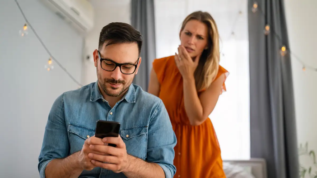 Affäre bei WhatsApp verstecken: Mann schaut lächelnd auf sein Handy, hinter ihm steht eine Frau