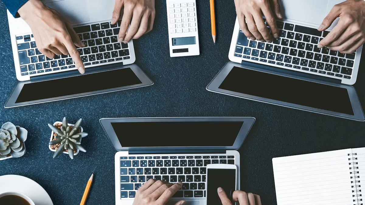 Vollzeitarbeit: Drei Laptops stehen aufgeklappt auf einem Tisch, an dem drei Personen arbeiten