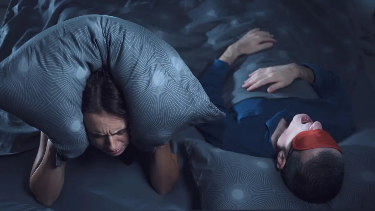 Frau zieht Kissen über den Kopf, weil Mann neben ihr im Schlaf spricht