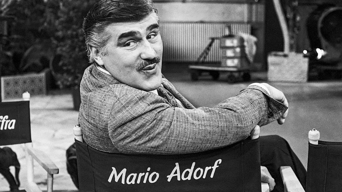 Mario Adorf schwarz-weiß 1988 auf einem Stuhl, auf dem sein Name steht