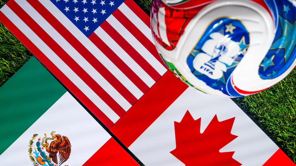 Fußball liegt neben Flaggen der Austragungsländer 2026 (USA, Mexiko und Kanada)