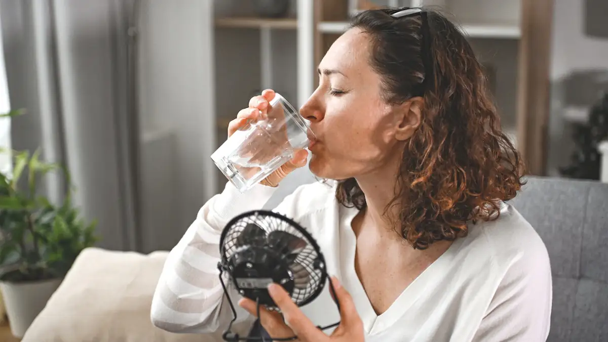 Frau trinkt bei Hitze Wasser, während sie einen kleinen Ventilator in der Hand hält