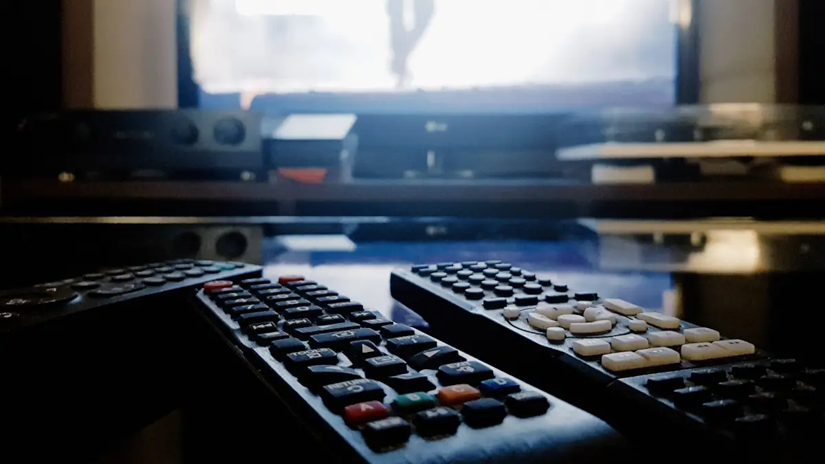 Nahaufnahme von zwei Fernbedienungen, im Hintergrund ein eingeschalteter Fernseher, der Strom verbraucht
