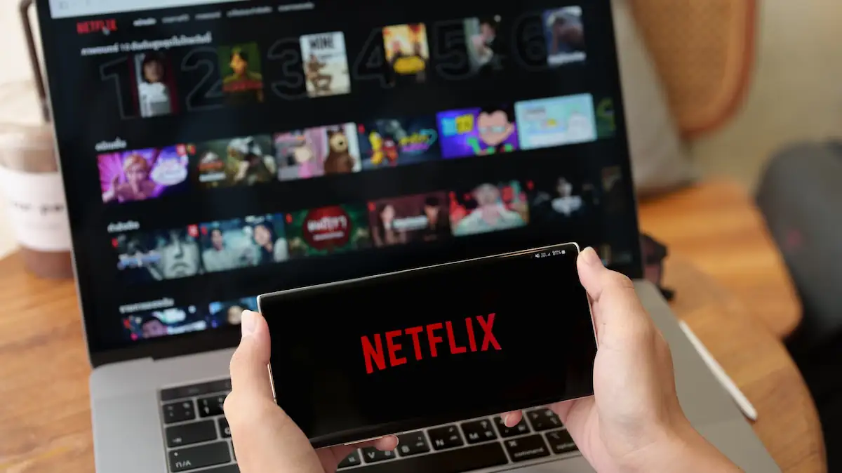Netflix-Logo auf einem Smartphone, das vor die Oberfläche des Streaming-Dienstes auf einem Laptop gehalten wird